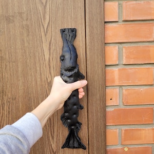 May include: Black metal door handle shaped like a fish. The handle is mounted on a wooden door.