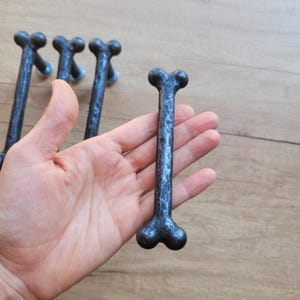 Può includere: Un set di pomelli per armadi a forma di osso, blu scuro. I pomelli hanno una superficie strutturata e sono tenuti in mano su uno sfondo di legno. I pomelli a forma di osso sono lunghi circa 15 cm.