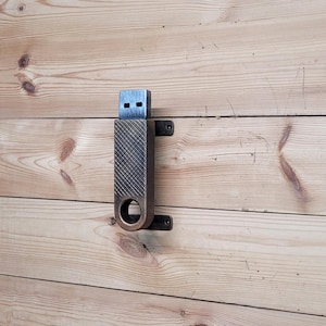 May include: A metal USB drive holder with a textured finish and a keyhole design. The holder is mounted to a wooden wall.
