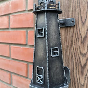May include: A black metal lighthouse door knocker with a weathered finish. The knocker is attached to a wooden door with a metal bracket.