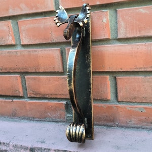 Puede incluir: Un llamador de puerta de metal negro y dorado con forma de cabeza de alce estilizada con un largo mango curvo. El llamador está montado en una pared de ladrillo.