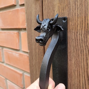 May include: Black metal door handle featuring a bull's head design. The handle is mounted on a wooden door and has a curved, ergonomic shape. The bull's head has detailed features, including horns and a textured surface.