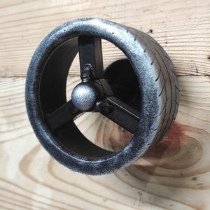 May include: A black metal wall hook shaped like a tire with a three-spoke design. The tire has a silver finish and a textured surface.