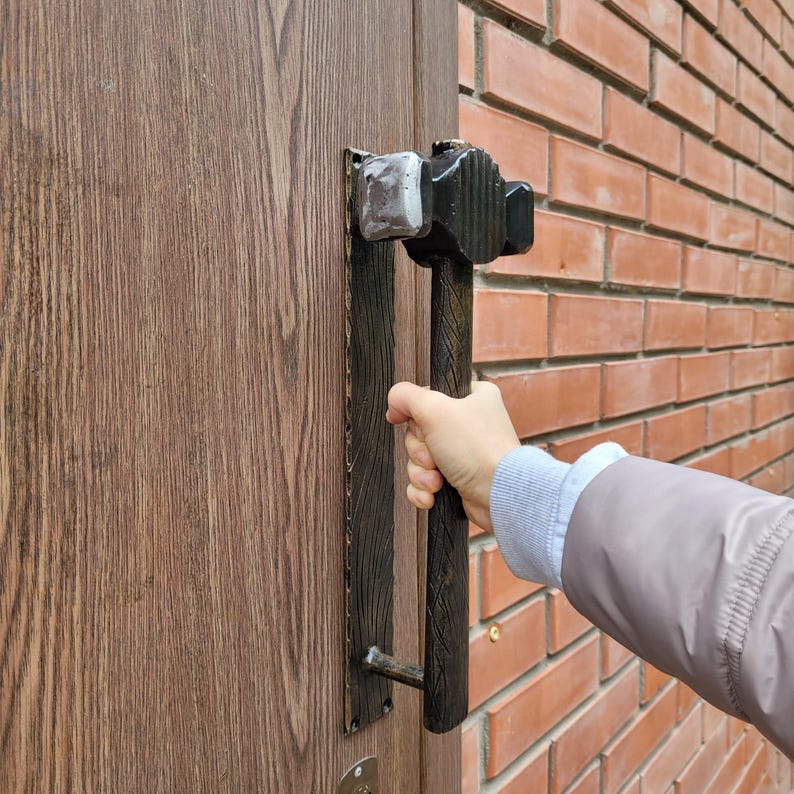 Puede incluir: Un tirador de puerta decorativo con forma de martillo. El tirador es negro y marr&oacute;n con una cabeza de martillo plateada. La puerta es de madera y la pared es de ladrillos rojos.