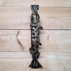 Könnte beinhalten: Ein bronzefarbener Türgriff in Fischform. Der Griff ist vertikal ausgerichtet und weist detaillierte Schuppen, Flossen und eine strukturierte Oberfläche auf. Der Griff ist auf einer hellen Holzoberfläche montiert.