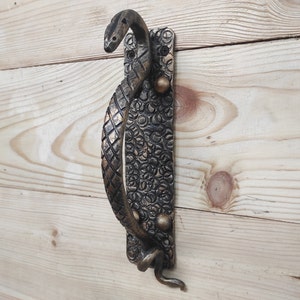May include: A decorative door handle shaped like a snake with a gold finish. The handle is mounted on a rectangular plate with a textured design.