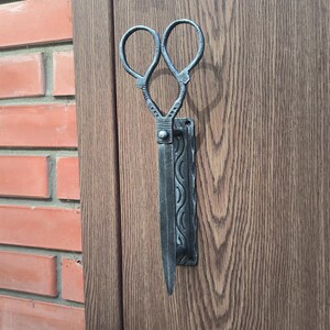 Op de afbeelding: Een zwarte metalen deurklopper in de vorm van een schaar. De deurklopper is gemonteerd op een houten deur.