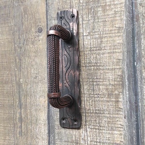 May include: A rustic door pull with a woven handle and a decorative backplate. The handle is made of a dark brown, rope-like material, and the backplate has an aged, textured finish. The door pull is mounted on a wooden door.