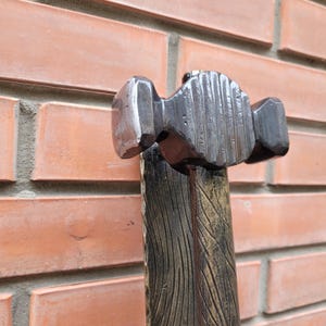 Puede incluir: Primer plano de un martillo de herrero. El cabezal del martillo es de metal oscuro y tiene una superficie texturizada. El mango de madera est&aacute; pintado con un dise&ntilde;o de vetas de madera. El fondo es una pared de ladrillos.