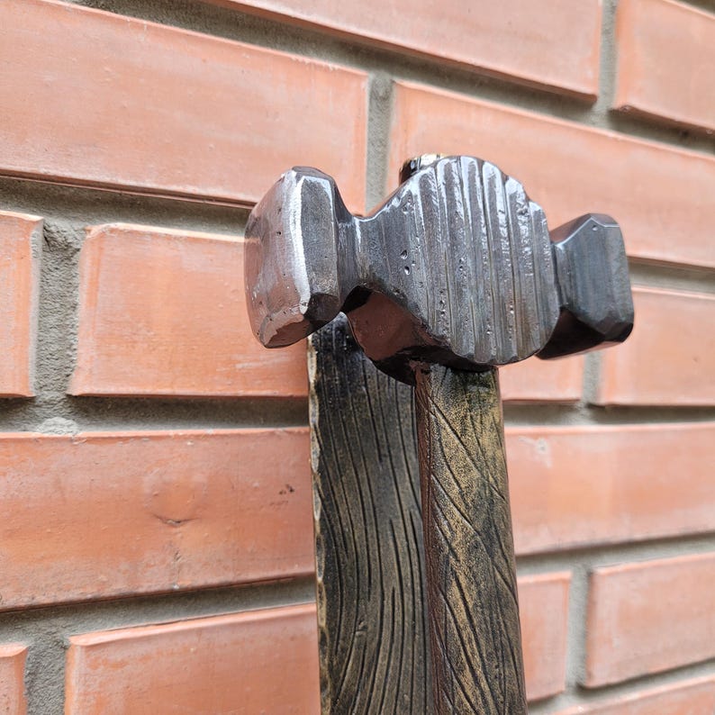 Puede incluir: Primer plano de un martillo de herrero. El cabezal del martillo es de metal oscuro y tiene una superficie texturizada. El mango de madera est&aacute; pintado con un dise&ntilde;o de vetas de madera. El fondo es una pared de ladrillos.