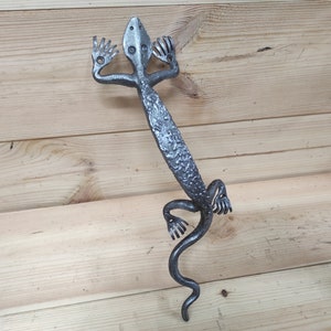 Op de afbeelding: Een zilveren metalen deurknop in de vorm van een hagedis met een textuur. De hagedis staat rechtop met zijn staart opgerold.