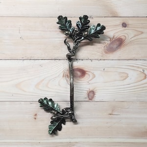May include: A metal hook shaped like an oak tree branch with leaves and acorns. The hook is designed to be mounted on a wall.