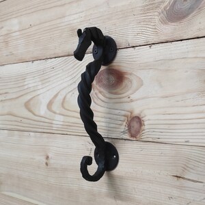 May include: Black metal door handle shaped like a seahorse with a twisted design. The handle is mounted on a wooden surface.