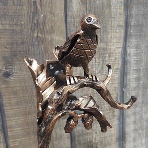 Könnte beinhalten: Bronze-farbene Metallvogelskulptur, die auf einem Ast mit Blättern und Eicheln sitzt. Der Vogel hat einen detaillierten, strukturierten Körper und ist auf einer dekorativen Halterung montiert. Die Skulptur ist für die Wandmontage konzipiert.