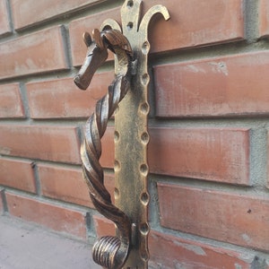May include: A gold-toned metal door handle with a twisted design and a horse head at the top. The handle is mounted on a brick wall.