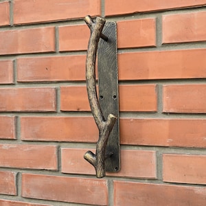 Könnte beinhalten: Ein rustikaler, goldfarbener, metallener Türgriff in Form eines Astes. Der Griff ist auf einer dunkelbraunen, hölzernen Rückplatte montiert. Der Griff ist an einer Backsteinwand befestigt.