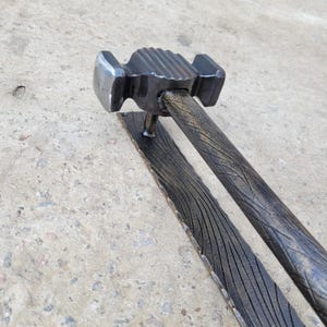 Puede incluir: Un martillo artesanal de metal oscuro con un mango texturizado y una base rectangular. La cabeza del martillo tiene una forma &uacute;nica con una parte superior estriada. El mango y la base tienen un patr&oacute;n de vetas de madera.