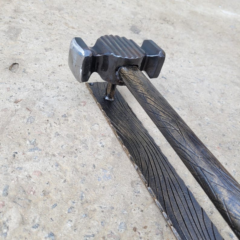 Puede incluir: Un martillo artesanal de metal oscuro con un mango texturizado y una base rectangular. La cabeza del martillo tiene una forma &uacute;nica con una parte superior estriada. El mango y la base tienen un patr&oacute;n de vetas de madera.