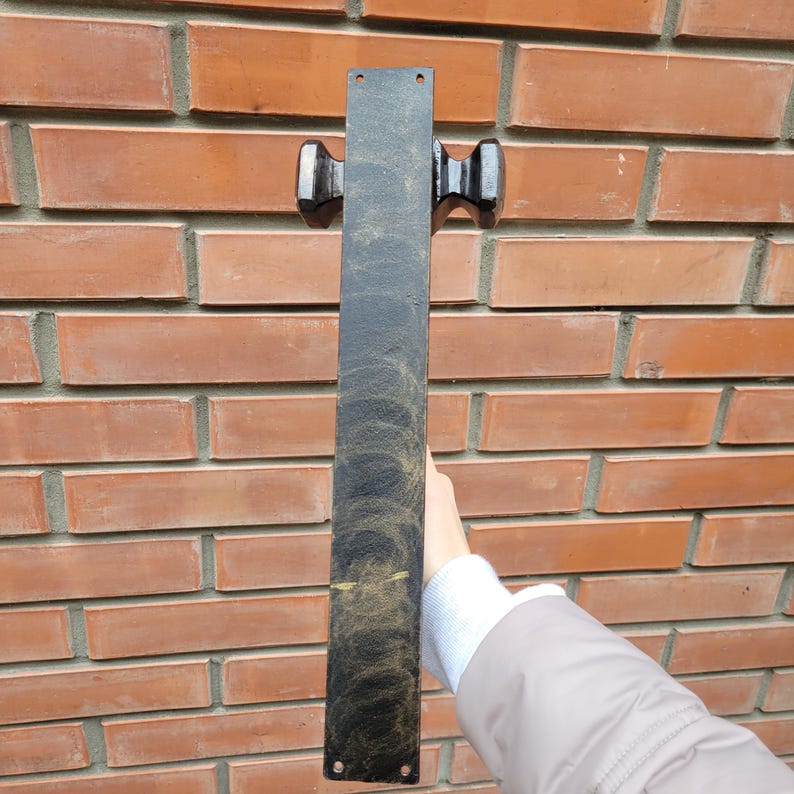 Puede incluir: Un tirador de puerta de metal negro con dos manijas decorativas. El tirador rectangular tiene una superficie texturizada con detalles dorados. El tirador de la puerta est&aacute; montado en una pared de ladrillo rojo.