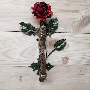 Op de afbeelding: Een rode metalen roos met groene bladeren en een gouden steel, bevestigd aan een houten deurknop.