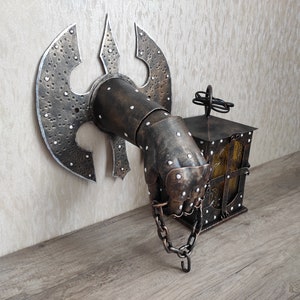 May include: A metal wall sconce in the shape of a hand holding a lantern. The hand is wearing a metal gauntlet with a large axe blade attached to the back. The lantern is made of metal and has a glass window. The sconce is painted in a dark brown colour with silver accents.
