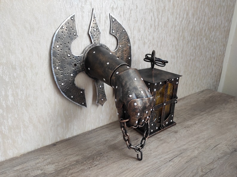 May include: A metal wall sconce in the shape of a hand holding a lantern. The hand is wearing a metal gauntlet with a large axe blade attached to the back. The lantern is made of metal and has a glass window. The sconce is painted in a dark brown colour with silver accents.
