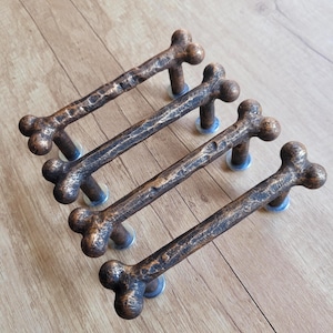 以下が含まれることがあります： ブロンズ色の骨型キャビネットプルが4つ。各プルはテクスチャ加工された表面を持ち、小さな丸い銀色のベースに取り付けられています。プルは、明るい茶色の木製の表面に斜めに配置されています。