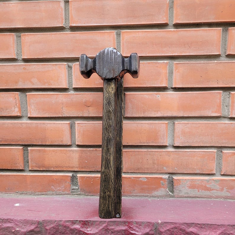 Puede incluir: Un martillo de metal con una cabeza oscura y texturizada y un mango de madera. El martillo est&aacute; colocado contra una pared de ladrillos rojos. El mango tiene un patr&oacute;n de vetas de madera.