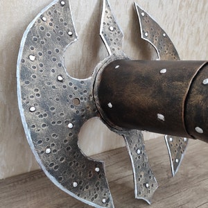 May include: A close-up of a metal wall decor piece with a textured, hammered finish. The piece is shaped like a stylised axe head with a circular centre and four pointed blades. The piece is painted in a dark brown colour with silver accents.