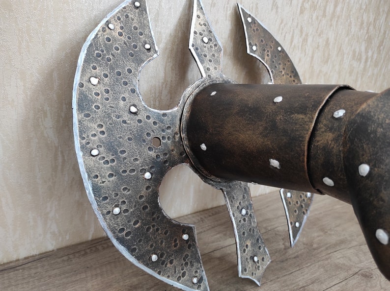 May include: A close-up of a metal wall decor piece with a textured, hammered finish. The piece is shaped like a stylised axe head with a circular centre and four pointed blades. The piece is painted in a dark brown colour with silver accents.