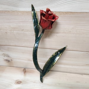 May include: A metal wall hook shaped like a red rose with green leaves. The hook is attached to a wooden surface.