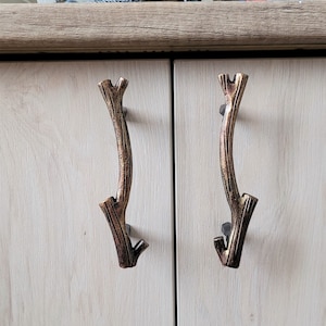 Puede incluir: Dos tiradores de puerta de armario con un diseño de rama. Los tiradores son de metal y tienen un acabado marrón oscuro.