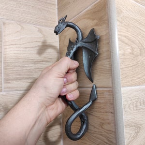 Op de afbeelding: Een zwarte metalen drakendeurgreep met een gekrulde staart en vleugels. De greep is gemonteerd op een muur en een hand houdt hem vast.