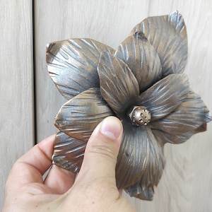 May include: Bronze-coloured, flower-shaped cabinet pull with detailed petal textures. The hardware features a central, textured knob. The floral design adds a decorative touch to furniture. The pull is held by a hand.