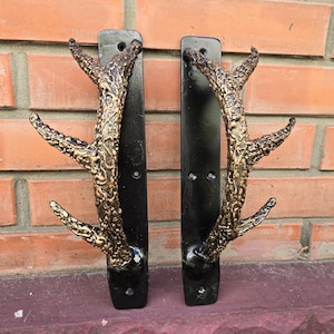 Könnte beinhalten: Zwei schwarze Metall-Türgriffe mit goldfarbenen Geweih. Die Griffe sind an einer Backsteinwand montiert.