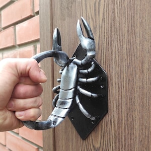 以下が含まれることがあります： 黒い金属製のベースに銀色のサソリのドアノッカー。サソリの尻尾は巻き上げられており、ノッカーは木製のドアに取り付けられています。