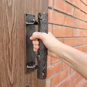 Peut inclure: Une poignée de porte rustique avec un design de branche d'arbre. La poignée est marron foncé et tenue par une main. La poignée est montée sur une plaque métallique foncée. La porte est en bois et le mur en briques rouges.