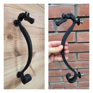 Op de afbeelding: Twee zwarte metalen deurgrepen in de vorm van een zeepaardje. De grepen zijn gemonteerd op een houten oppervlak en een bakstenen muur.