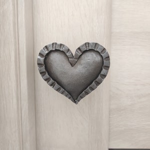May include: A heart-shaped metal door handle with a textured, scalloped edge. The handle is a dark gray color and is mounted on a light wood door.