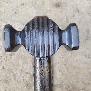 Puede incluir: Primer plano de un martillo de metal oscuro con una cabeza texturizada y acanalada y un mango estampado. La cabeza del martillo tiene una forma redondeada y sim&eacute;trica con dos extremos ensanchados. El mango tiene un dise&ntilde;o retorcido.