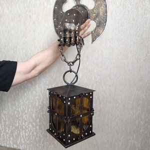 May include: A metal wall sconce with a hand-shaped design holding a lantern. The lantern is made of metal and has a glass panel. The sconce is attached to a wall with a chain.