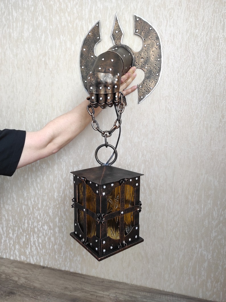 May include: A metal wall sconce with a hand-shaped design holding a lantern. The lantern is made of metal and has a glass panel. The sconce is attached to a wall with a chain.