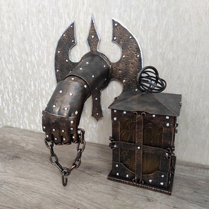 May include: A wall-mounted metal lamp with a hand-shaped base and a lantern-style shade. The base is decorated with rivets and a chain. The lantern has a glass panel and a metal frame.