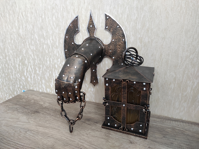 May include: A wall-mounted metal lamp with a hand-shaped base and a lantern-style shade. The base is decorated with rivets and a chain. The lantern has a glass panel and a metal frame.
