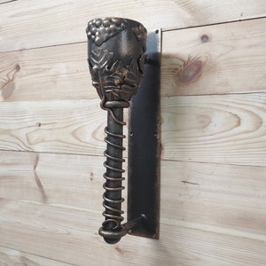 May include: A wall-mounted wine bottle holder with a decorative metal design featuring grapes and leaves. The holder is made of dark metal and has a spiral design.