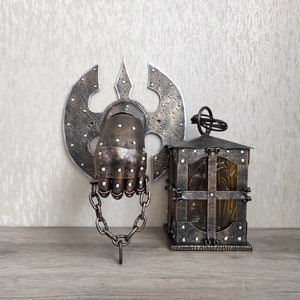 May include: A metal wall decoration featuring a hand in a gauntlet holding a chain, with a decorative axe behind it. A small, metal lantern with a glass window sits on a wooden surface.