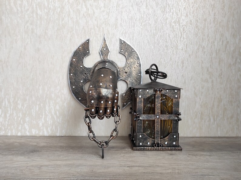 May include: A metal wall decoration featuring a hand in a gauntlet holding a chain, with a decorative axe behind it. A small, metal lantern with a glass window sits on a wooden surface.