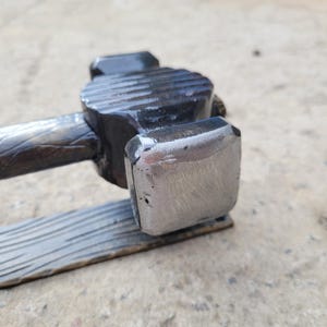 Puede incluir: Primer plano de un martillo de metal forjado a mano. La cabeza del martillo es de metal oscuro y texturizado con una cara de golpeo rectangular. El mango es de metal oscuro y texturizado. El martillo est&aacute; dise&ntilde;ado para uso intensivo.