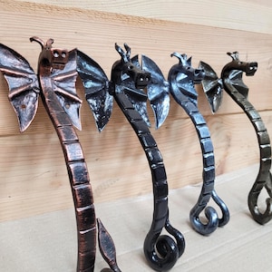 Puede incluir: Cuatro manijas de puerta de metal con forma de dragón hechas a mano en tonos bronce, negro y gris. Cada manija presenta una cabeza de dragón con alas detalladas y un cuerpo curvado. Las manijas están montadas sobre madera.