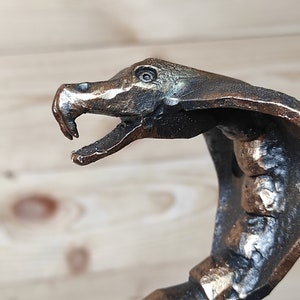 May include: A metal cobra-shaped bottle opener with a dark bronze finish. The head of the cobra is raised and the mouth is open, revealing sharp teeth. The body of the cobra is curled around the bottle opener.